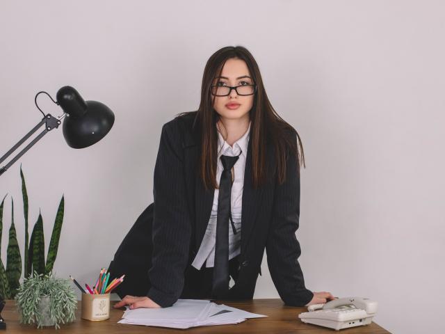 ChristinaBliss, con lunghi capelli castani, occhiali e un completo a righe, è appoggiata a una scrivania, mostrando il seno, ed è circondata da matite, una lampada e un telefono bianco..