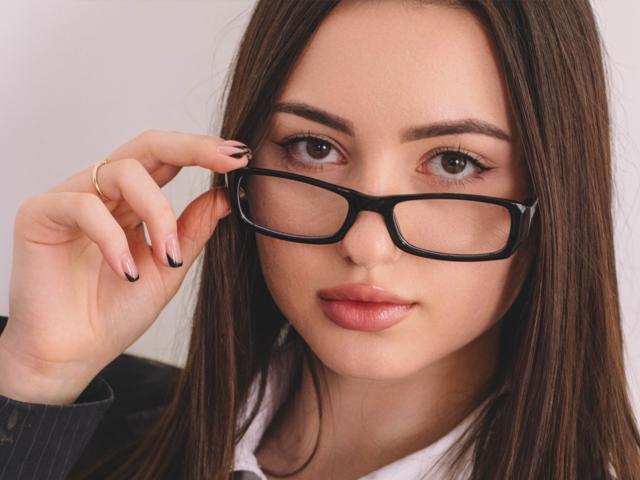 ChristinaBliss, con lunghi capelli castani, indossa occhiali neri, ha una mano vicino al viso, mostrando un anello d'oro, e ha un'espressione seria con rossetto rosa..