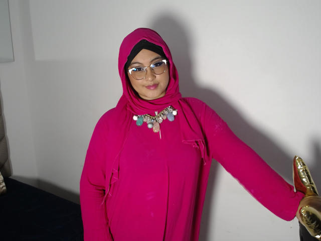 HelueJamil vestindo um hijab rosa, óculos e um vestido rosa com um colar de prata, segurando uma estátua dourada com a mão direita, sorrindo levemente..