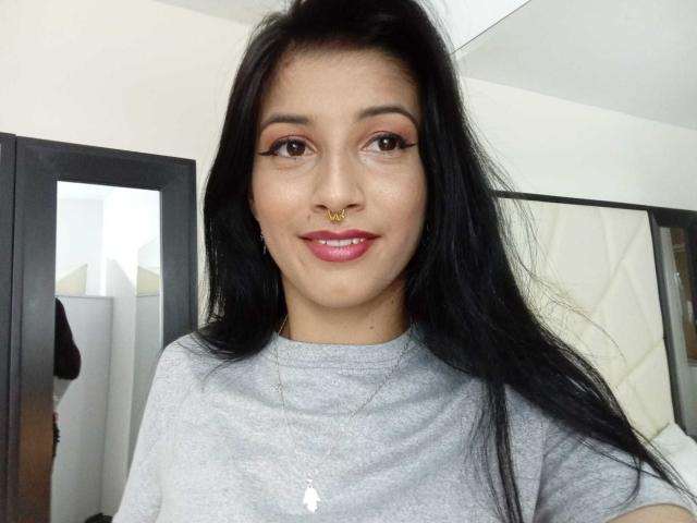 AlisKooper has long black hair, brown eyes, pink eyeliner, nose ring, and gray shirt, smiling with pink lipstick, wearing a gold necklace with a small white pendant.
