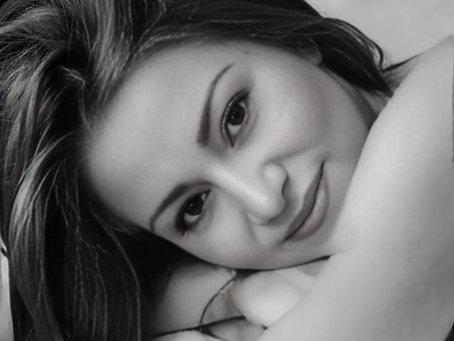 KatePri has her head resting on her arm, smiling softly at the camera. Her dark, wavy hair is spread out, and she's wearing a subtle, relaxed expression.