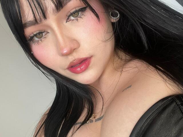 DanielaRuizz has pale skin, green eyes, black hair, pink blush, red lipstick, small nose, black eyeliner, earrings, tattoo on right chest, black top, slight smirk, soft light.