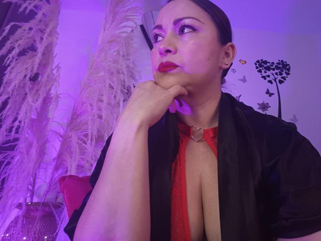 KatePri with dark hair, red lipstick, and black open shirt, showing cleavage, resting chin on hand, wearing heart choker, in purple-lit room with puffy white plant.