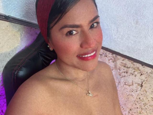 IvannaDuran, topless, with red headscarf, smiling, showing her tits, wearing a necklace with a small heart pendant, and red lipstick, sitting on a black leather chair.
