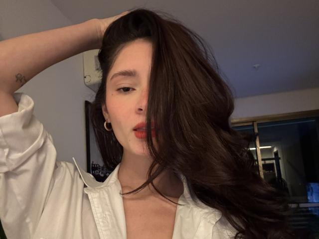 Fefezinha with long brown hair covering one eye, wearing a white shirt, hand in hair, small tattoo on arm, gold hoop earrings, slightly open mouth, soft light.