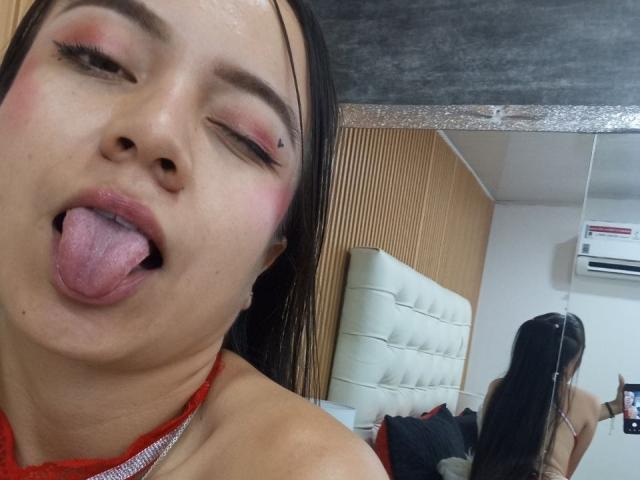 JessyLenon with her tongue out, eyes half-closed, pink eyeshadow, red lace top, mirror reflection, wet black hair, small nose piercing, and a slightly sweaty face.