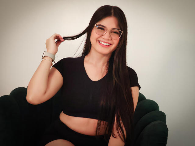 CamilaSofiia with long black hair, smiling, wearing glasses, black crop top, and black shorts, holding hair with right hand, sitting on green chair, wearing white smartwatch.