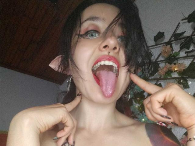 MeiJaisen with black hair, green eyes, nose piercing, red lipstick, tongue out, fingers pointing at face, visible tattoo on shoulder, wearing earrings, looking at camera.