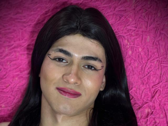 MichellPratTrans has dark brown hair, thick eyebrows, eyeliner wings, and pink lipstick. Her skin is light with a slight sheen. Her face is centered against a bright pink background.
