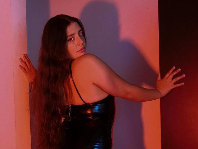 Paola Lyon, con lunghi capelli castani, indossa un abito in lattice nero lucido, guarda oltre la spalla, con le mani appoggiate alle pareti, illuminazione rossa e rosa, ombra sul muro, carina, sexy..