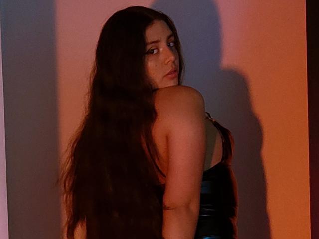 PaolaLyon with long dark hair, looking over her shoulder, wearing a black strapless leather dress, shadow on the wall, subtle makeup, slight pout, curvy with a visible butt.