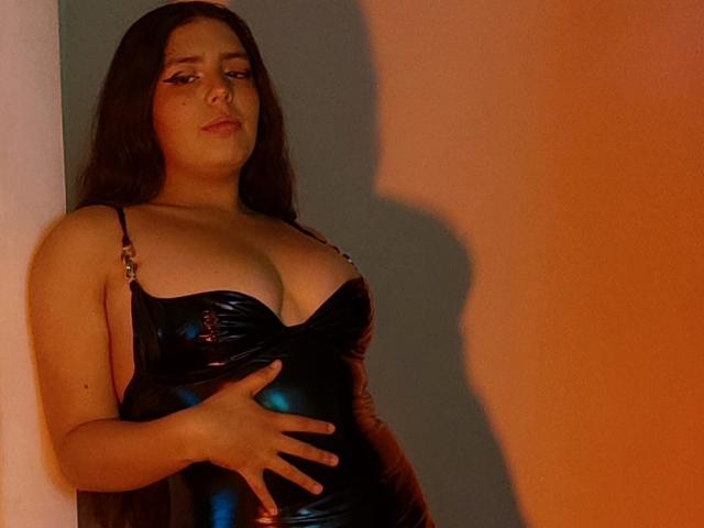 PaolaLyon with long dark hair, wearing a shiny black latex dress, hand on her belly, shadow on wall, looking slightly annoyed. Her tits are prominent, she's curvy.