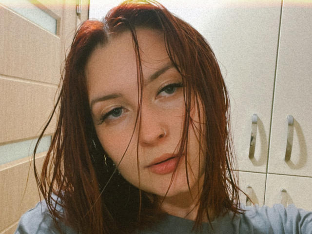 DarkXTemptress has wet brown hair covering her face, light makeup, and a nose ring, wearing a gray shirt, with a slight, relaxed expression in a soft-lit, indoor setting.