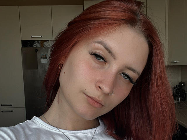 Red-haired DarkXTemptress with pale skin, small nose, and subtle smile, wearing white shirt, has small tits, visible nose ring, and subtle makeup, in a kitchen setting.