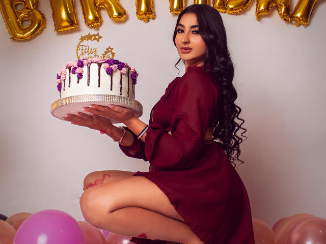 JasmineRh with long black hair, wearing a red sheer dress, holding a white cake with purple flowers and chocolate drips, kneeling on pink balloons, showing her tatted thigh.