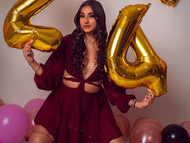 JasmineRh with long dark curly hair, wearing a deep red, cut-out dress, holding shiny gold "4" balloons, surrounded by pink and black balloons, sitting on pink balloons.