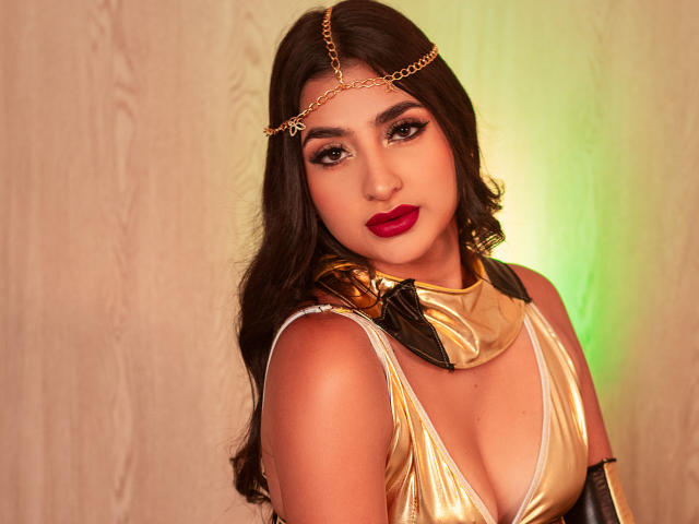 JasmineRh with dark hair, gold head chain, and red lipstick, wearing a shiny gold top that reveals her tits, has a confident, sexy look. Her makeup is bold.