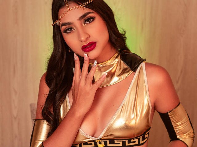 JasmineRh with dark hair, red lipstick, gold chain headpiece, metallic gold top showing tits, gold arm cuffs, and long nails, touching her face, looking at the camera.