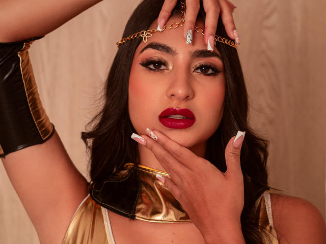 JasmineRh has dark hair, gold headpiece, red lipstick, and long nails. She's wearing a gold and black top with a black arm cuff, touching her face.
