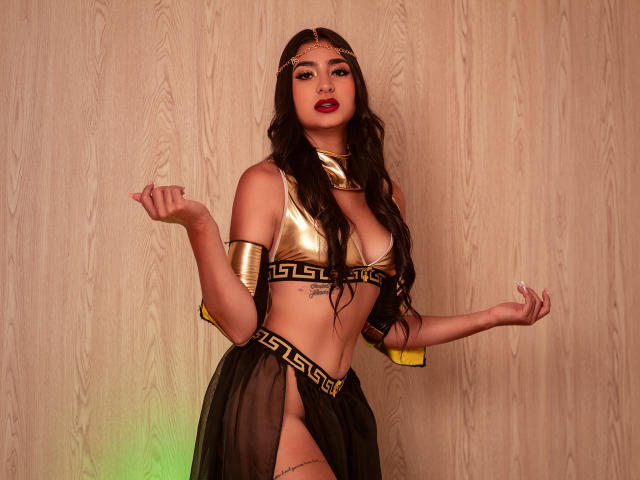 JasmineRh with long dark hair, gold headpiece, gold bra, black Grecian-style skirt, gold arm cuffs, and red lipstick, standing with hands up, showing her tits and ass.