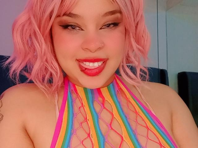 NatieRush, com cabelo rosa ondulado, piercing no nariz e batom vermelho, mostrando a língua, vestindo um top de rede colorido com alças, exibindo seus seios. Sua maquiagem é marcante, com delineador..
