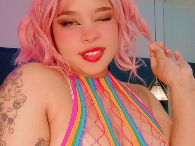 NatieRush, avec ses cheveux roses, clignant de l'œil, portant un haut en filet arc-en-ciel, montrant ses seins, avec du rouge à lèvres, un anneau au nez et des tatouages sur son bras gauche, souriant avec la langue tirée..