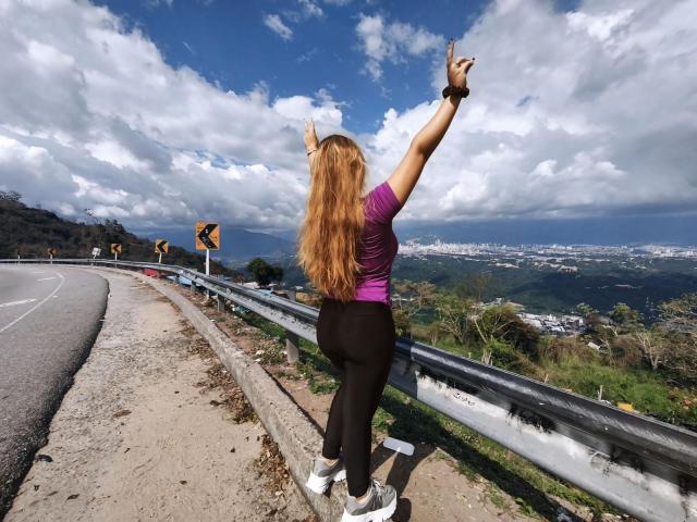 ZoeTobon69 stands with arms raised, pointing to the sky. She has long red hair, a purple shirt, black pants, and white sneakers, on a curvy road with metal railings.