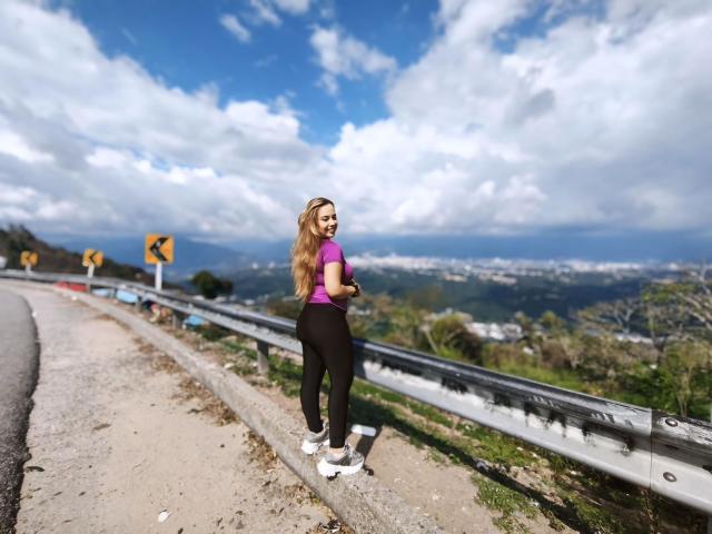 ZoeTobon69 standing on a gravel road, wearing a purple top, black ass-hugging pants, and white sneakers, with long blonde hair, looking back at the camera.