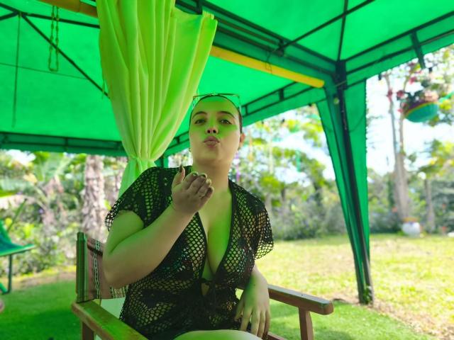 ZoeTobon69 sitting under a green canopy, wearing a black mesh top that reveals her tits, with green curtains, holding a cigarette, looking to the side, in a grassy area.