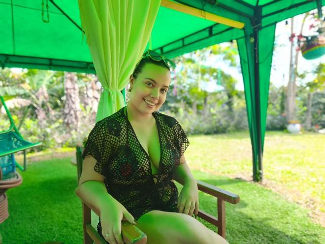 ZoeTobon69 sitting under a green canopy, wearing a black mesh top with visible tits, smiling, green grass, and a wooden chair, with a sun hat on her head.