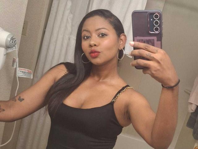DianaReine with dark skin and long black hair, wearing a black tank top showing her tits, red lipstick, hoop earrings, and holding a phone, taking a mirror selfie.