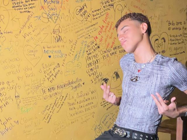 DavidLomas, with curly brown hair and a cute face, wearing a striped shirt and black belt, is writing on a yellow wall with a pen, looking happy and relaxed.