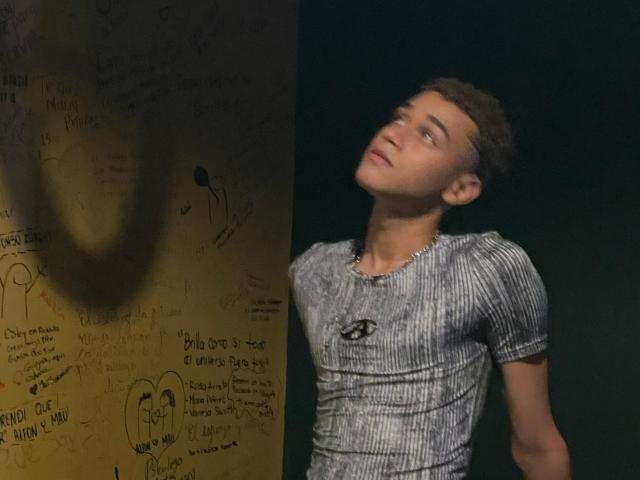 DavidLomas with short brown hair, wearing a tight gray striped shirt, leaning against a wall with graffiti, looking upward with a slightly confused expression.