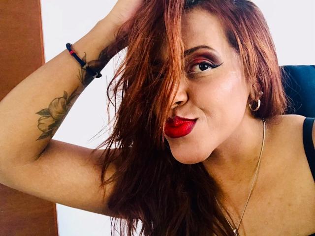 ShamanthaFire has red lipstick, a tattoo on her arm, and wavy brown hair covering one eye. She's smiling with a hand in her hair, wearing a black top.