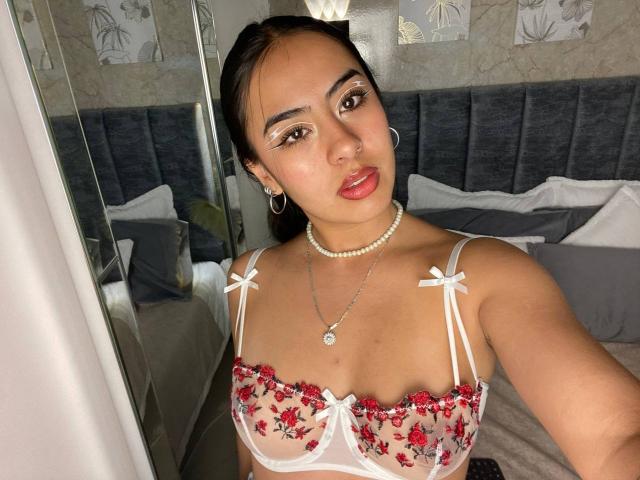 SamanthaRosses with tan skin, dark hair, red lipstick, wearing a sheer red floral bra, pearl necklace, and hoop earrings, showing her boobs in a bedroom selfie.