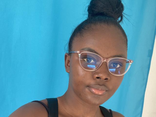 LuciCrees, avec une peau foncée, un haut noir, des lunettes et un chignon haut, souriant légèrement, sur un fond bleu, avec des lèvres brillantes et un reflet subtil dans ses yeux..