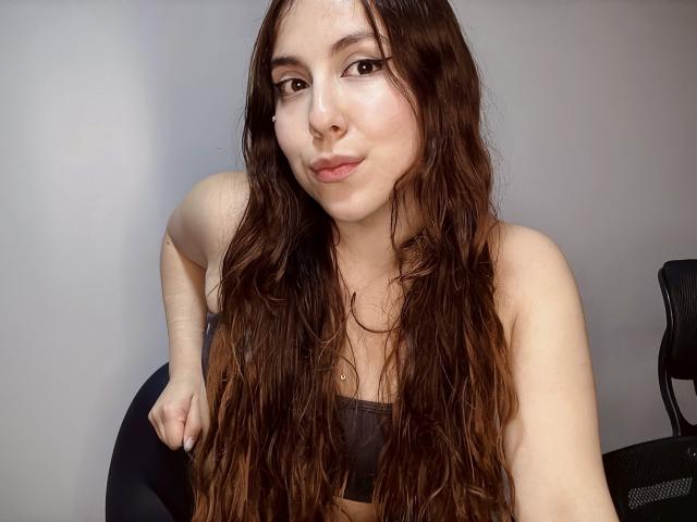 AmberlStha, con lunghi capelli castani ondulati, indossa un reggiseno sportivo grigio, ha un carino piercing al naso ed è seduta su una sedia nera, sorridendo leggermente alla telecamera..