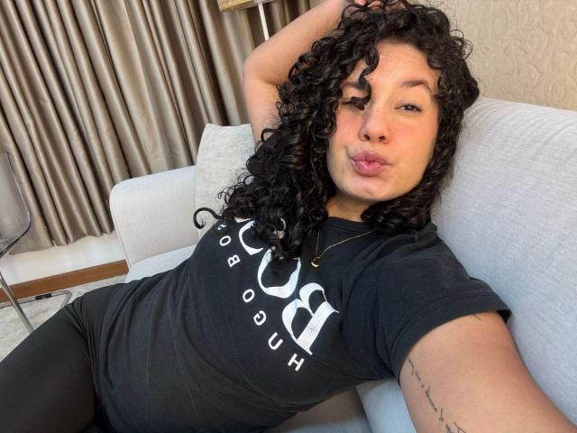 RachellBeaumont with curly black hair, pouting lips, wearing a black T-shirt, black pants, and a necklace, lounging on a gray couch, her arm tattoo visible.