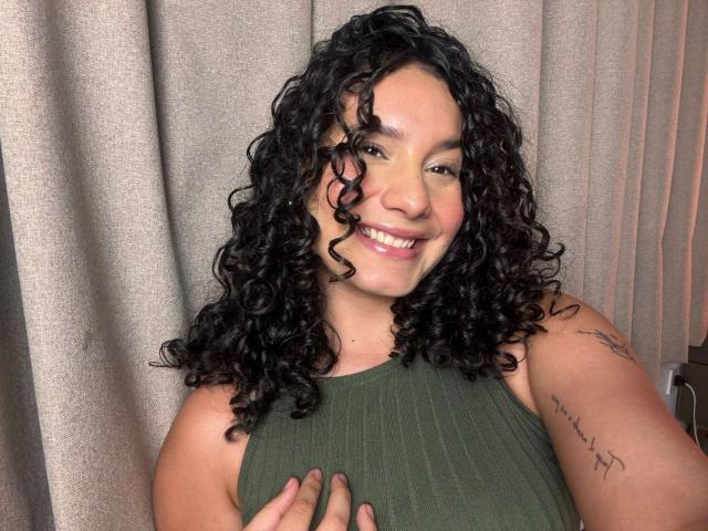 RachellBeaumont with curly black hair, smiling, wearing a green ribbed tank top, hand on chest, tattoo on right arm, light brown skin, and pink lipstick.