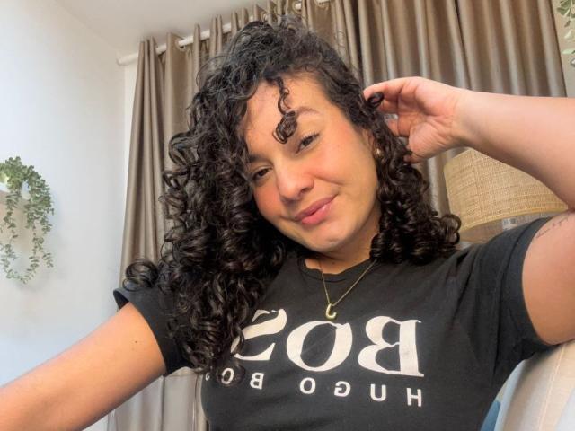 RachellBeaumont with curly black hair, brown skin, small nose ring, black t-shirt, subtle smile, right hand in hair, gold necklace, and tattoo on right arm.