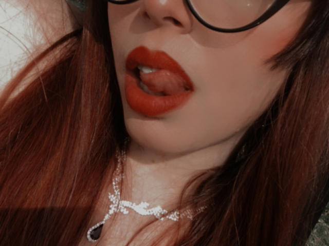 Rosalinnes with red lipstick, tongue out, wearing glasses, and a silver necklace with a black pendant, showing her pouty face and reddish-brown hair.