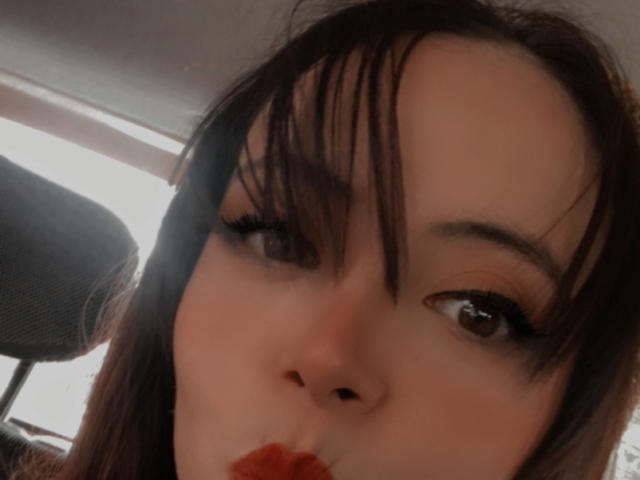 Rosalinnnes' face, brown eyes, long black eyelashes, dark brown hair with bangs, sucking on red lipstick, soft light, subtle smile, slightly blurred, close-up selfie.