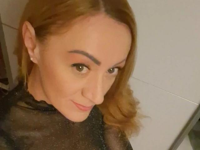 Irina-Faust-hot with brown hair, thick eyebrows, subtle makeup, pink lipstick, and a black sheer top, smiling slightly at the camera. Her nose is centered, and she's wearing small earrings.