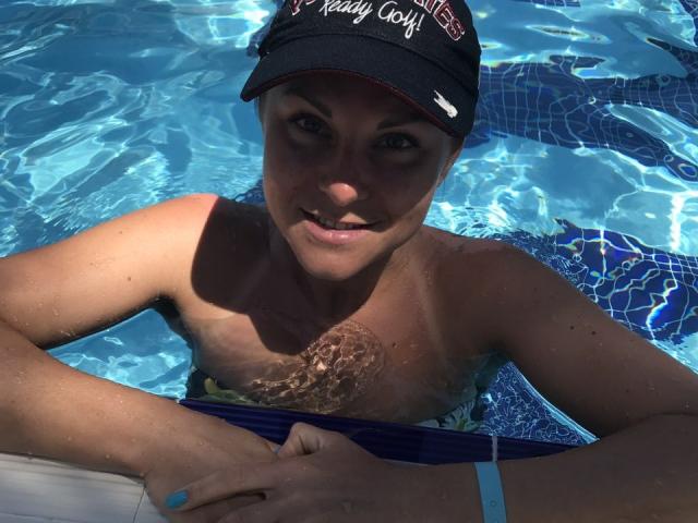 BelleLisaG in a pool, wearing a black cap, blue wristband, and swim trunks, with wet skin and a smile, hands on a white pool float. Blue water reflects sunlight.
