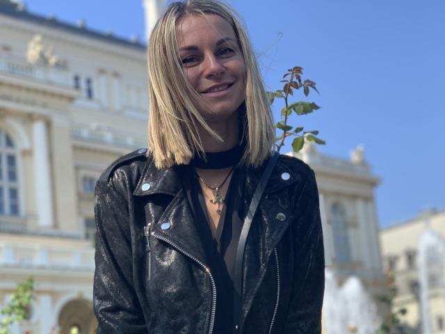 BelleLisaG with blonde bob, black leather jacket, black choker, and necklace, smiling, holding a plant, blue sky, and ornate building in background.