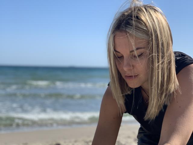 BelleLisaG with blonde hair, looking down, has a serious expression, wearing a black shirt, on a sandy beach with gentle waves in the background.