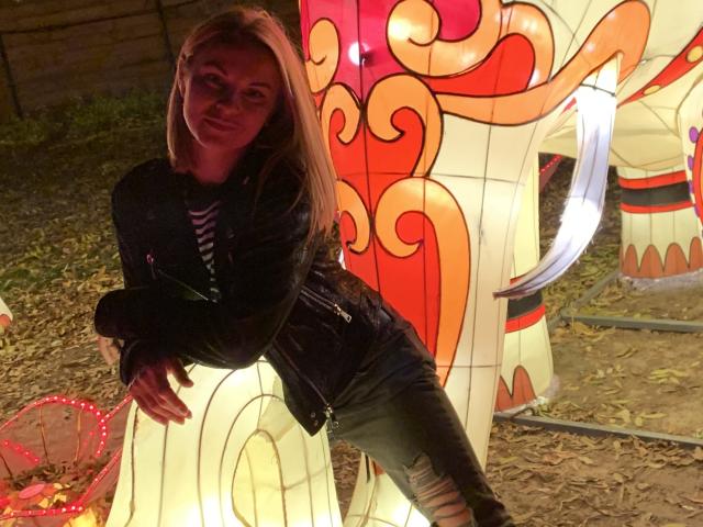 BelleLisaG with blonde hair, wearing a black jacket and ripped jeans, leaning on a brightly lit, colorful, inflatable dragon at night. Her tits are partially visible.