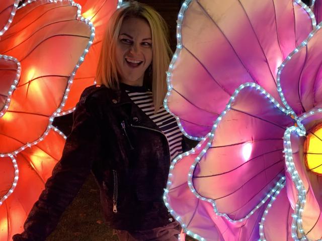 BelleLisaG with blonde hair, smiling, wearing a black leather jacket and striped shirt, standing in front of large, glowing, pink and orange flower lanterns.