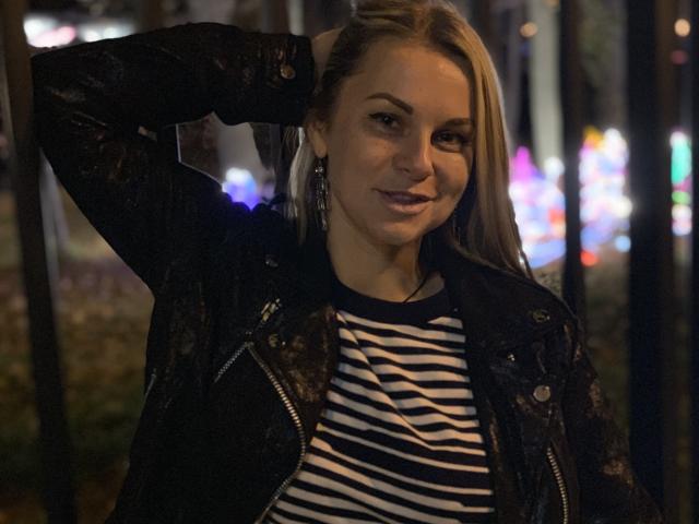 BelleLisaG with blonde hair, black lace jacket, striped shirt, and earrings, hand in hair, smiling, dark lighting, bokeh background, visible nipples under shirt.