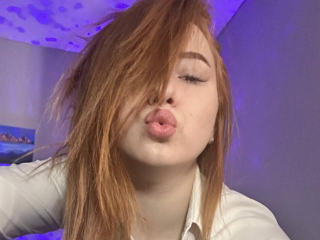 Barberry with red hair, pouty pink lips, and long eyelashes, kissing camera with eyes closed, wearing white shirt, in dim purple-lit room.
